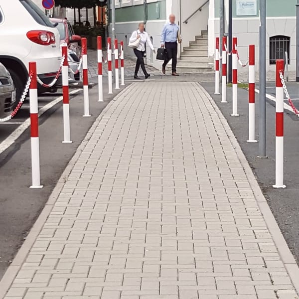 Gepflasterter Gehweg mit MORION-Sperrpfosten 60 rot-weiß, zwei Fußgänger, Treppe zum Gebäude; Autos links geparkt.