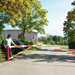 Eine Person hebt eine COMPACT Gatterschranke am Parkplatz-Eingang an, im Hintergrund stehen Bäume und ein Auto.