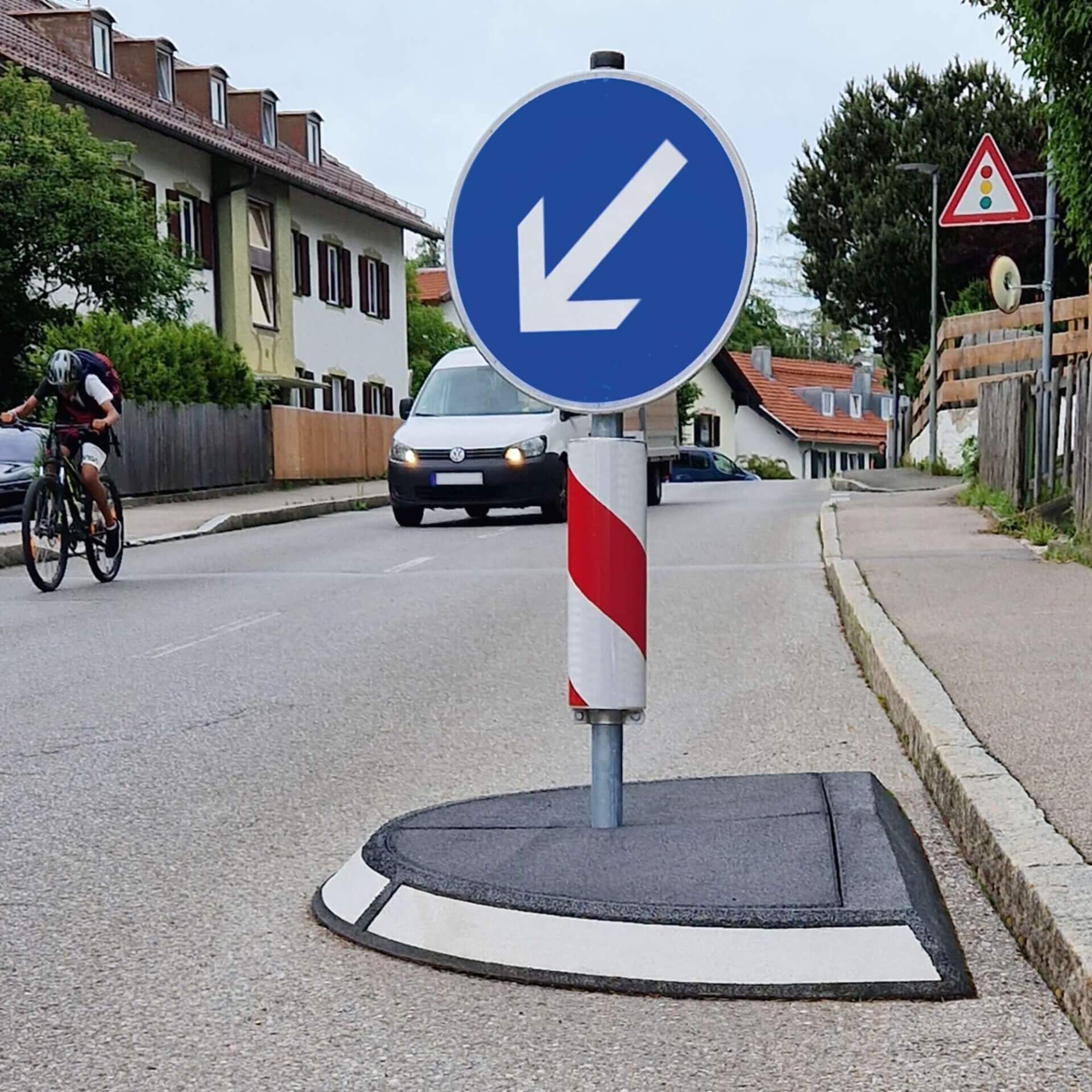 GAMMA Verkehrsinsel mit blauem Verkehrsschild, weißem Linkspfeil, Radfahrer und Auto im Hintergrund, Straße in Deutschland.