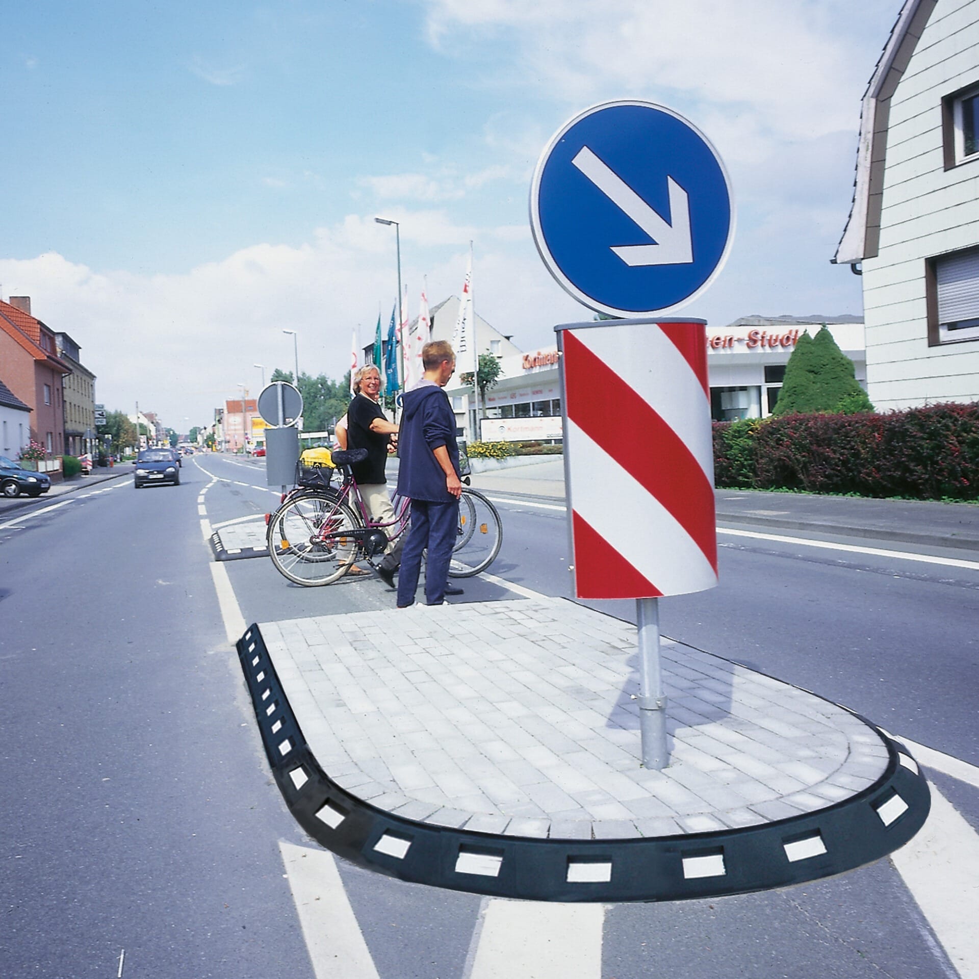 Zwei Personen mit Fahrrädern warten auf Verkehrsinsel mit GAMMA Elastikbordstein, umgeben von Verkehrs- und Warnschildern, Gebäude im Hintergrund.