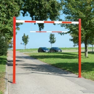 Roter und weißer Durchfahrtbegrenzer aus Metall auf gepflasterter Straße, zwei parkende Autos, Bäume im Hintergrund.