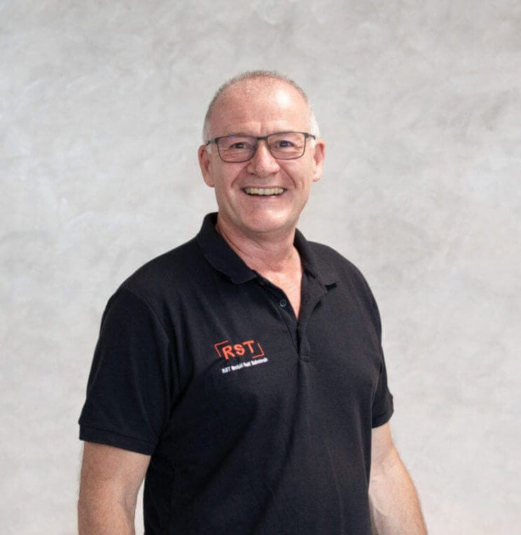 Mann mit kurzem grauem Haar lächelt, trägt dunkles Hemd vor hellem Hintergrund; Porträt für Technische Industrieberatung.