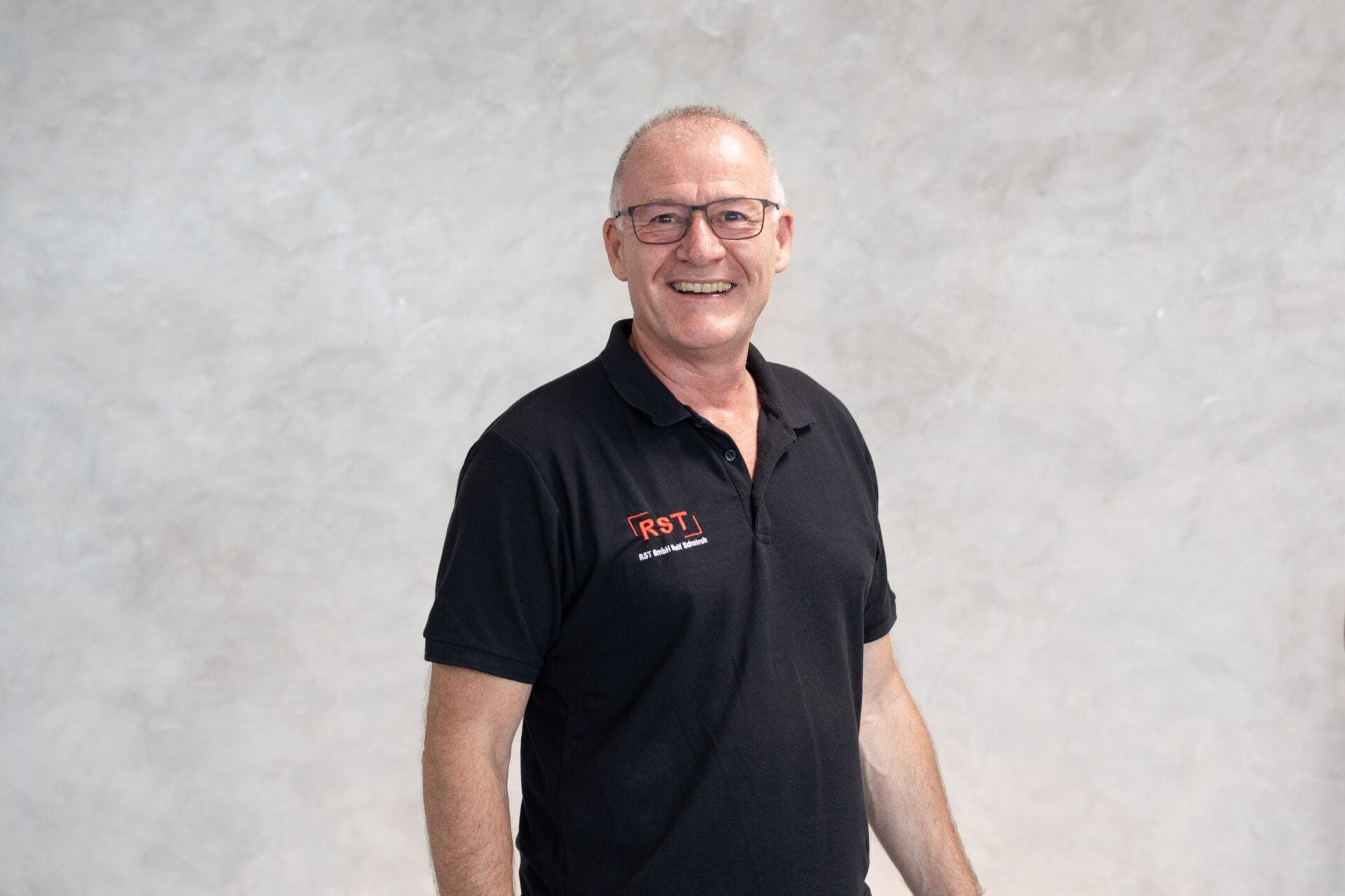 Mann mit kurzem grauem Haar lächelt, trägt dunkles Hemd vor hellem Hintergrund; Porträt für Technische Industrieberatung.