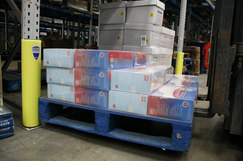 Blaue Palette mit zahlreichen Plastik-umwickelten Kartons steht im Lagergang neben Metallregalen und gelben Regalstützschutz.