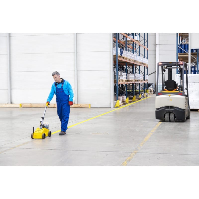 Ein Mann in blauer Arbeitskleidung markiert Lagerhallenboden mit Verkehrsmarkierungsset; Gabelstapler steht daneben.