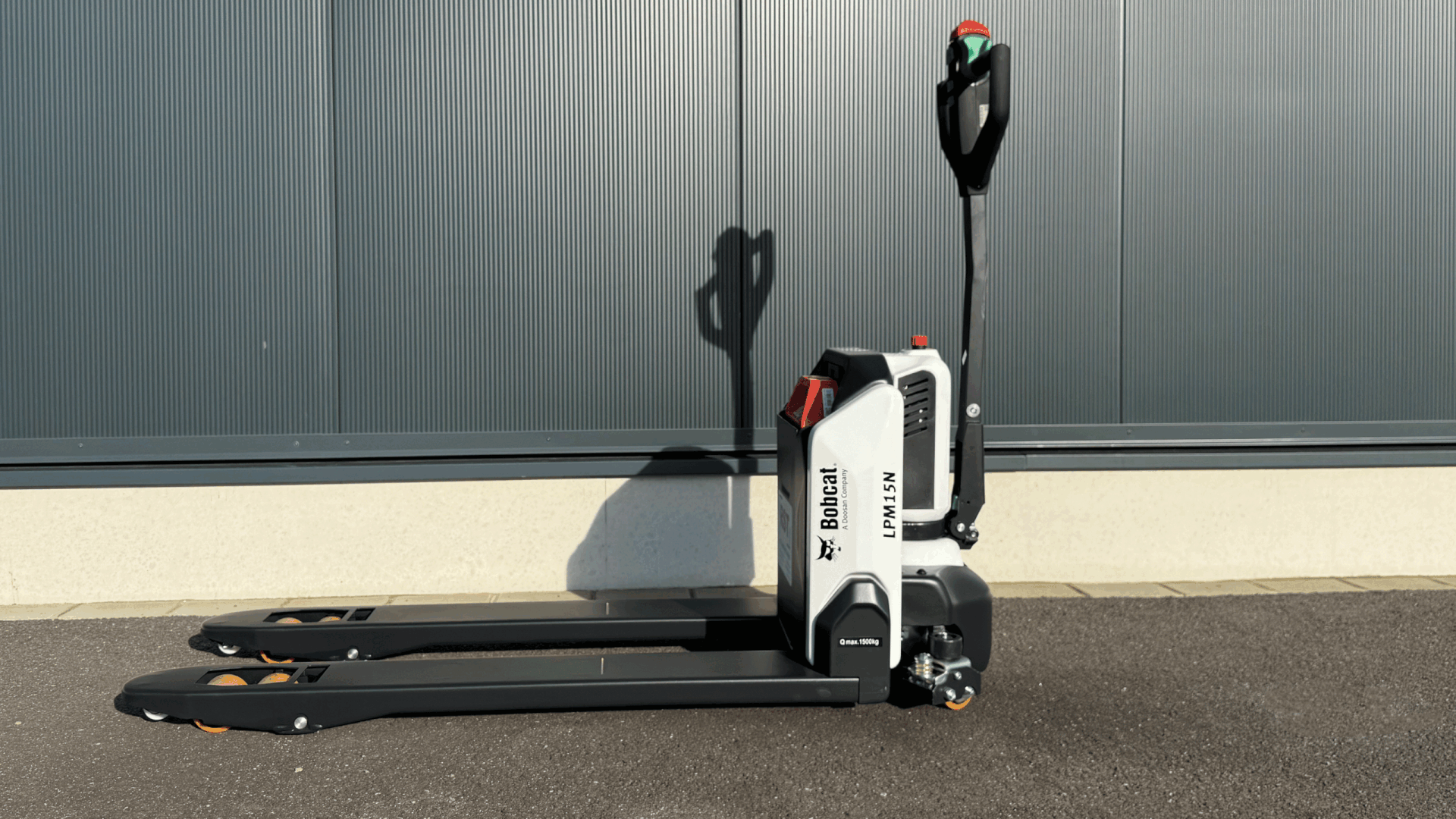 Ein Bobcat Elektrohubwagen steht auf Asphalt vor einer grauen Industriehalle, Griff senkrecht, Gabeln nach vorn ausgestreckt.