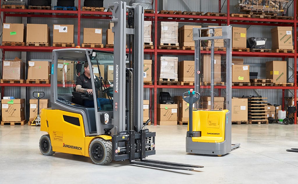 Ein Mann fährt einen gelben Gabelstapler im Lager; daneben steht ein unbemannter Palettenheber, Regale mit Kartons im Hintergrund.