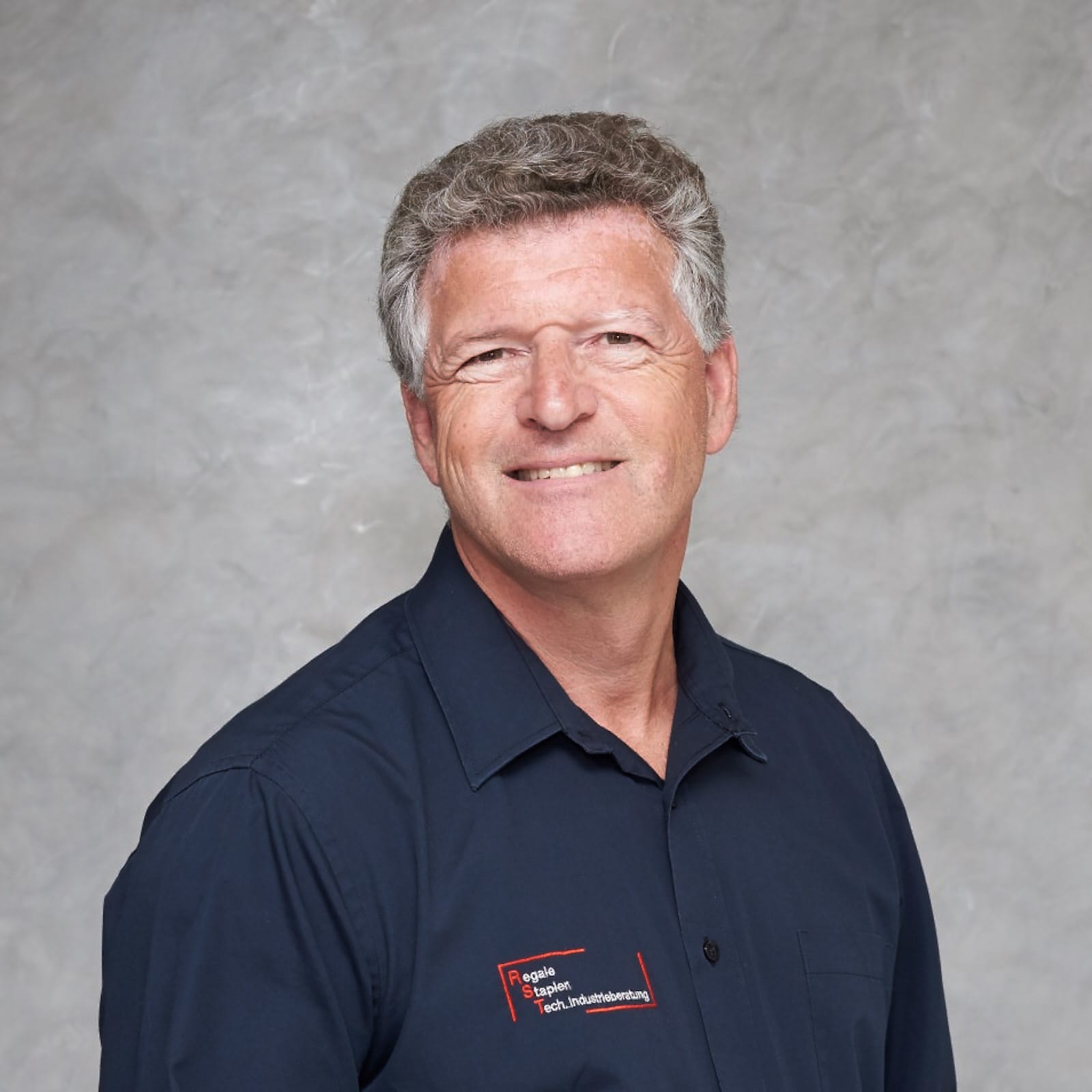 Mann mit kurzem grauem Haar lächelt, trägt dunkles Hemd vor hellem Hintergrund; Porträt für Technische Industrieberatung.