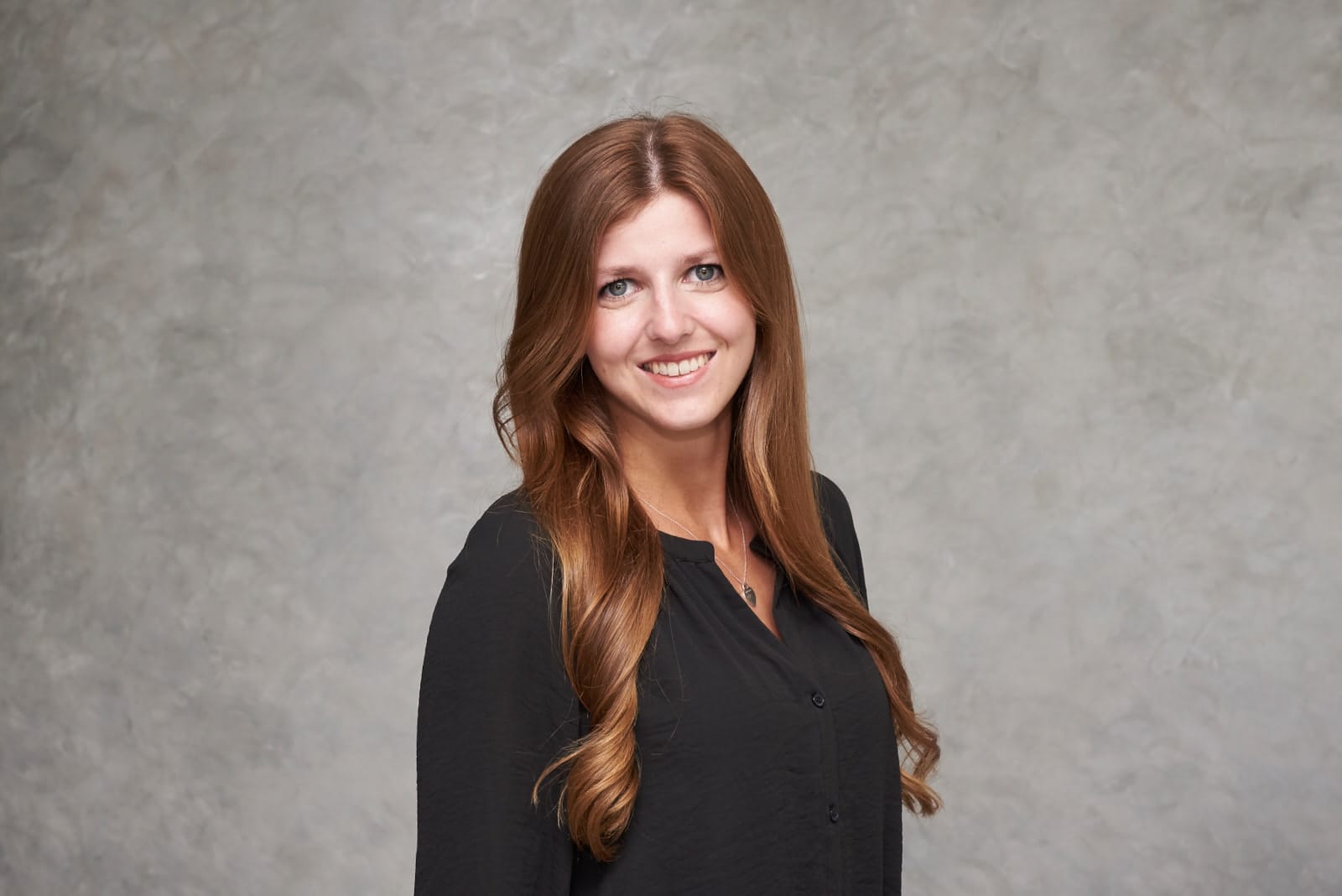 Junge Frau mit langen, glatten, hellbraunen Haaren im schwarzen Oberteil lächelt frontal vor hellem grauem Hintergrund.