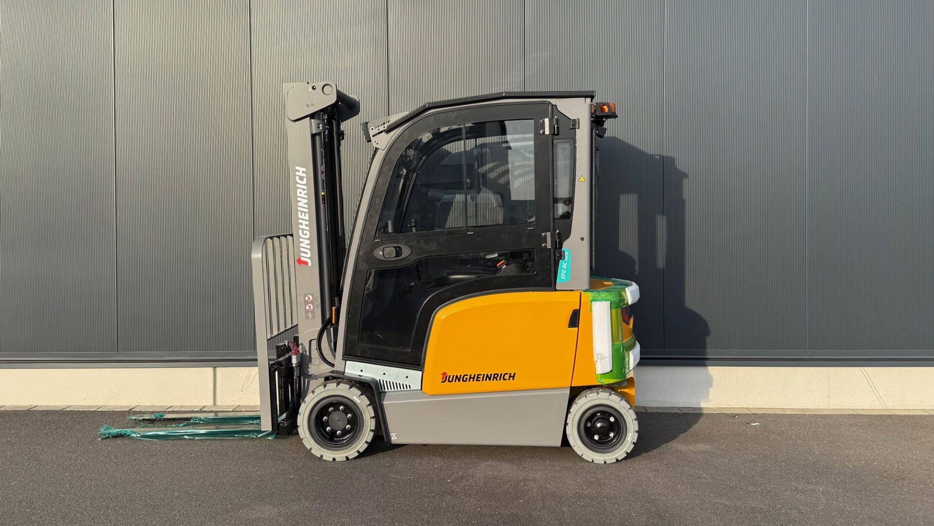 Jungheinrich Elektro-Gabelstapler steht auf Asphalt vor einer grauen Wand. Industrie-Fahrzeug, Lagerlogistik, Seitensicht.