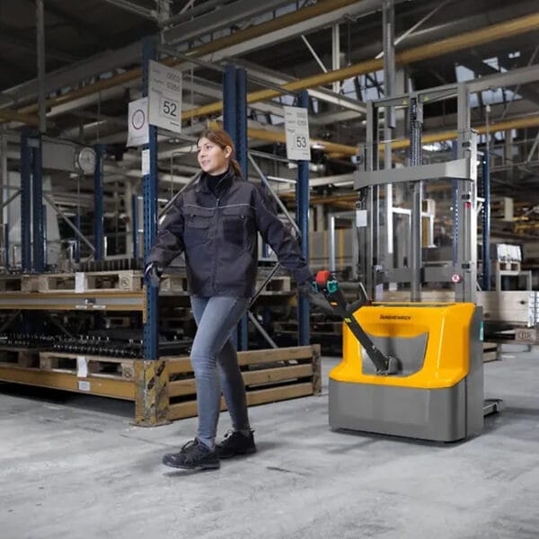 Frau in Arbeitskleidung bedient Elektro-Deichselhochhubwagen AMC 12 (280ZT) im Lager vor Regalen mit gelagerten Materialien.