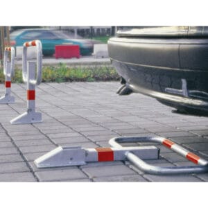 Metall Kipp-Pfosten mit roten Reflexstreifen sichern einen asphaltierten Parkplatz, Auto geparkt, weitere Fahrzeuge unscharf im Hintergrund.