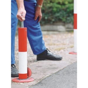 Person hebt rot-weißen PARATlift Absperrpfosten vom gepflasterten Gehweg am Straßenrand, trägt blaue Hose und schwarze Schuhe.