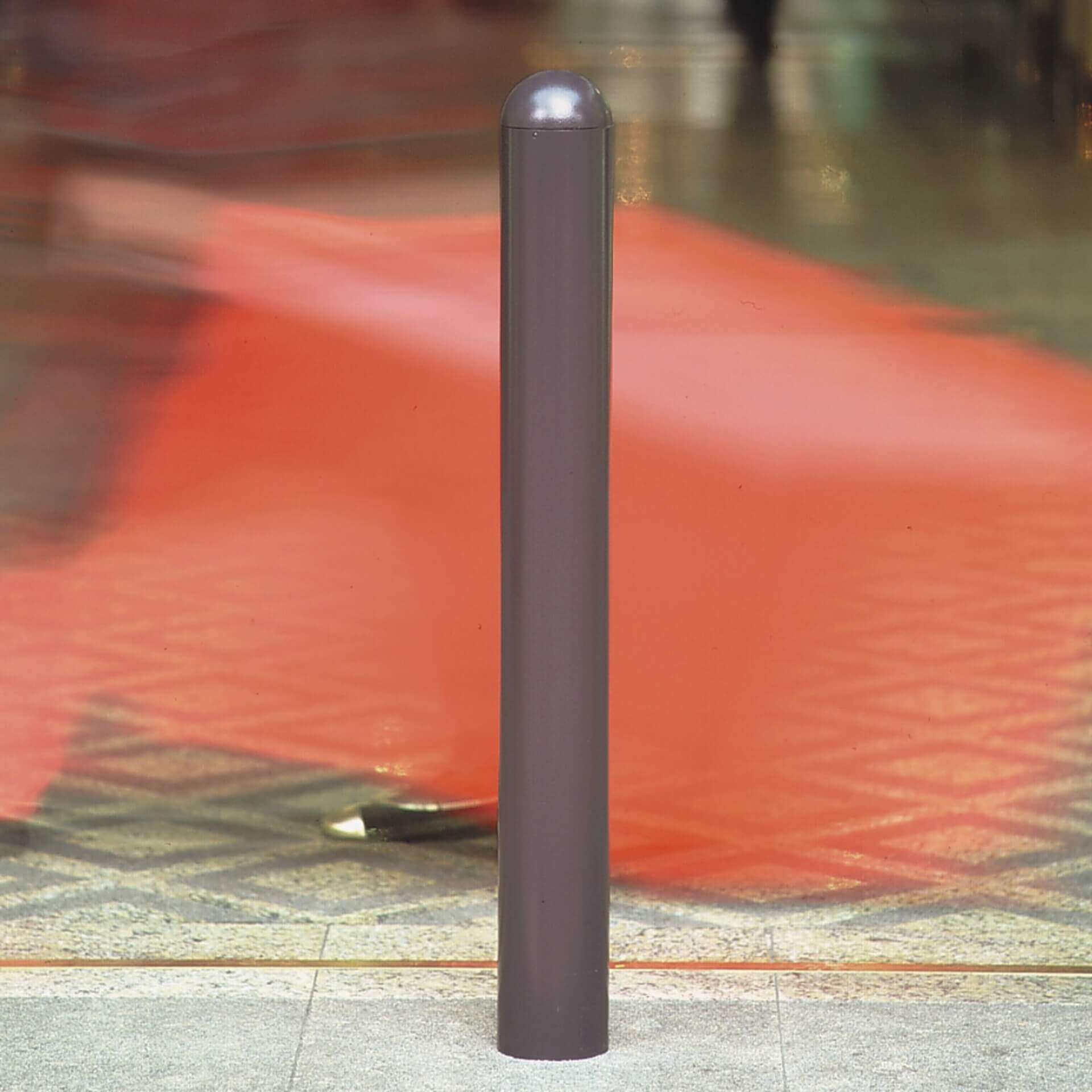 Grauer City-Poller WIEN 90 aus Metall, zylindrisch, steht auf Fliesen; unscharfer Hintergrund mit roten und braunen Passanten.