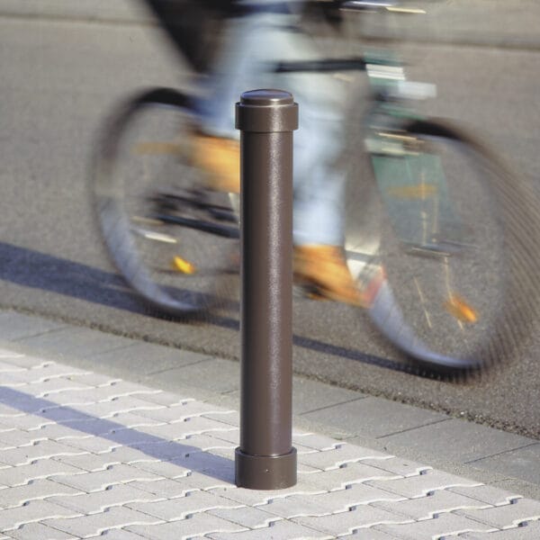 Brauner Metall-Stadtpoller ZÜRICH auf gepflastertem Gehweg, im Hintergrund fährt ein Radfahrer vorbei.