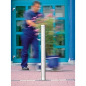 Edelstahlpoller MILANO auf gefliestem Gehweg, unscharfe Person schiebt Pflanze im Topf vor blauen Türen im Hintergrund.