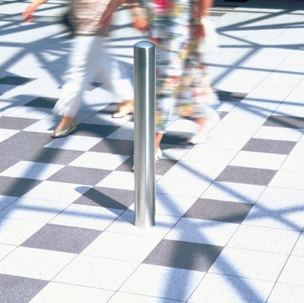 Zwei unscharfe Passanten gehen am Edelstahlpoller BERLIN auf kariertem Fliesenboden, Sonnenlicht wirft lange Schatten.