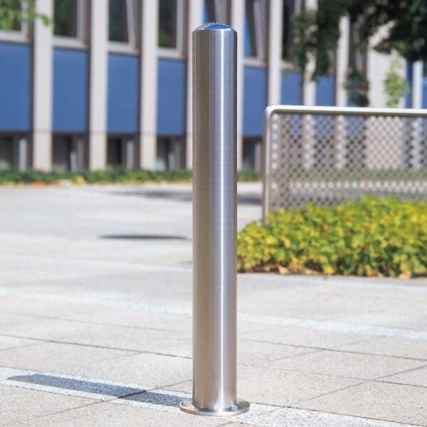 Edelstahlpoller BERLIN auf gefliestem Außenboden, im Hintergrund Gebäude und Grünanlage sichtbar.