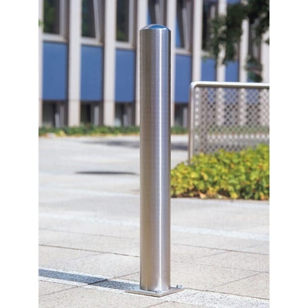 Edelstahlpoller BERLIN auf gepflastertem Gehweg vor Gebäude mit blauen und beigen Wänden, Grünpflanzen im Hintergrund.