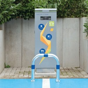 CITY-Schutzbügel GROß an E-Ladestation mit blauen und gelben Markierungen, umgeben von Grünfläche und Betonbarrieren.