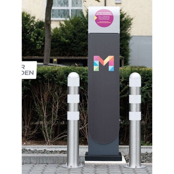 Ladestation für Elektroautos mit buntem M-Logo, zwei Stahl-Schutzpollern, Pflasterfläche, Büschen und Gebäuden im Hintergrund.