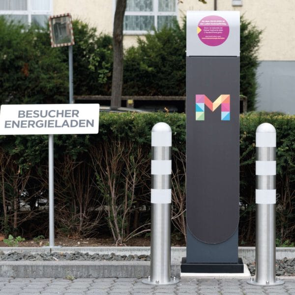 CITY-Schutzpoller aus Edelstahl an E-Ladestation mit buntem M-Logo und Schild "Besucher Energieladen" auf dem Parkplatz.