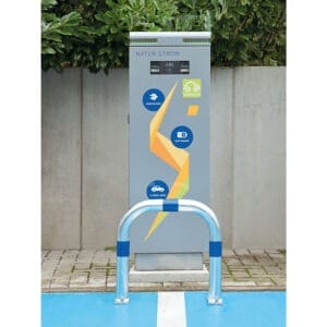 CITY-Schutzbügel GROß aus Metall vor Natur Strom E-Ladestation auf gepflastertem Platz, umgeben von Grün und Betonwand.