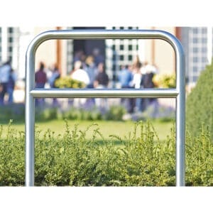 CITY-Anlehnbügel SPRINT aus Metall vor grünen Büschen, unscharfe Personengruppe und Gebäude im Hintergrund sichtbar.