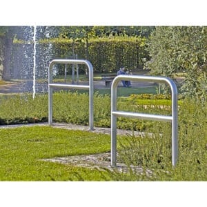 Zwei CITY-Anlehnbügel SPRINT Fahrradständer aus Metall auf Wiese im Park, mit Brunnen und sitzender Person auf Bank im Hintergrund.