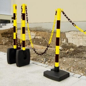 Drei gelb-schwarz gestreifte Ketten-Warnständer mit Kunststoffketten sperren eine Baustelle neben einem Gebäude ab.