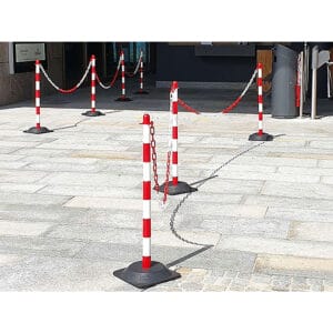 Sechs Kettenständer mit rot-weißen Streifen, durch Ketten verbunden, bilden eine Absperrung auf dem Gehweg vor einem Eingang.