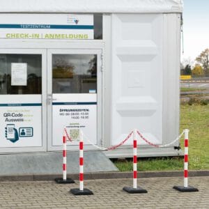 Weißes Testzentrum mit Glastür Check-in/Anmeldung, Öffnungszeiten, QR-Code Schild und GUARDA-Plus Ketten-Warnständer in Rot-Weiß.
