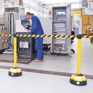 Arbeiter in blauer Arbeitskleidung an offenem Schaltschrank, vorn gelber-schwarzer MORION Gurt-Warnständer, Industrieumgebung.
