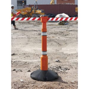 Absperrpfosten mit ausziehbarem orangem Gurt und rot-weißem Band sichern Baustelle; Bauarbeiter und Maschinen im Hintergrund.