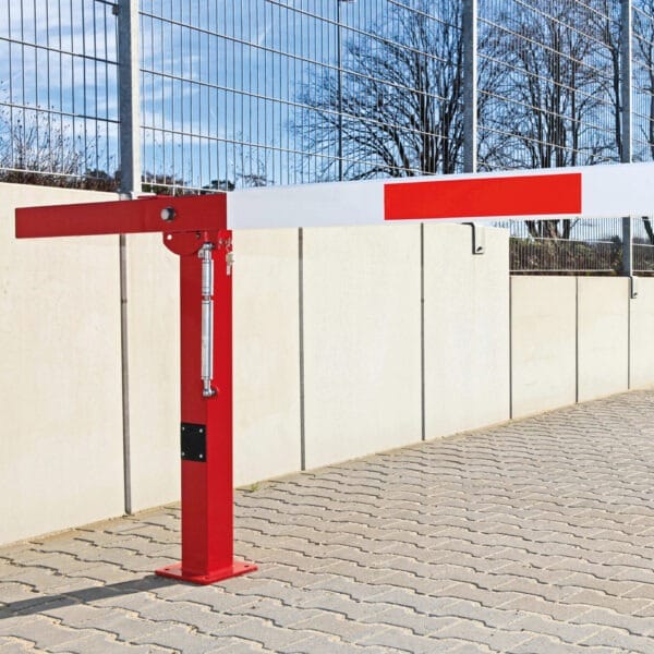 Rote und weiße COMPACT Handschranke steht auf Pflaster neben hohem Metallzaun und Betonwand, Absperrung Parkplatz Tor.