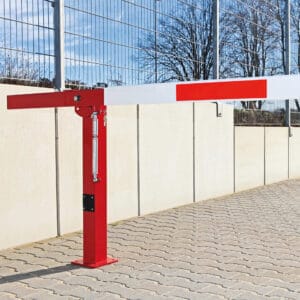 Rote und weiße COMPACT Handschranke aus Metall auf Asphalt neben Betonmauer und Maschendrahtzaun, teils bewölkter Himmel.