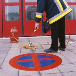 Person in Uniform markiert mit Gasbrenner ein rot-blaues Halteverbotsschild auf dem Asphalt vor roten Türen, Gasflasche dabei.
