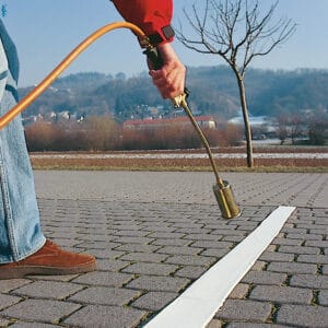 Person erhitzt PROline-Thermoplastik-Markierungslinie mit Gasbrenner auf Asphalt, Baum und Hügel im Hintergrund.