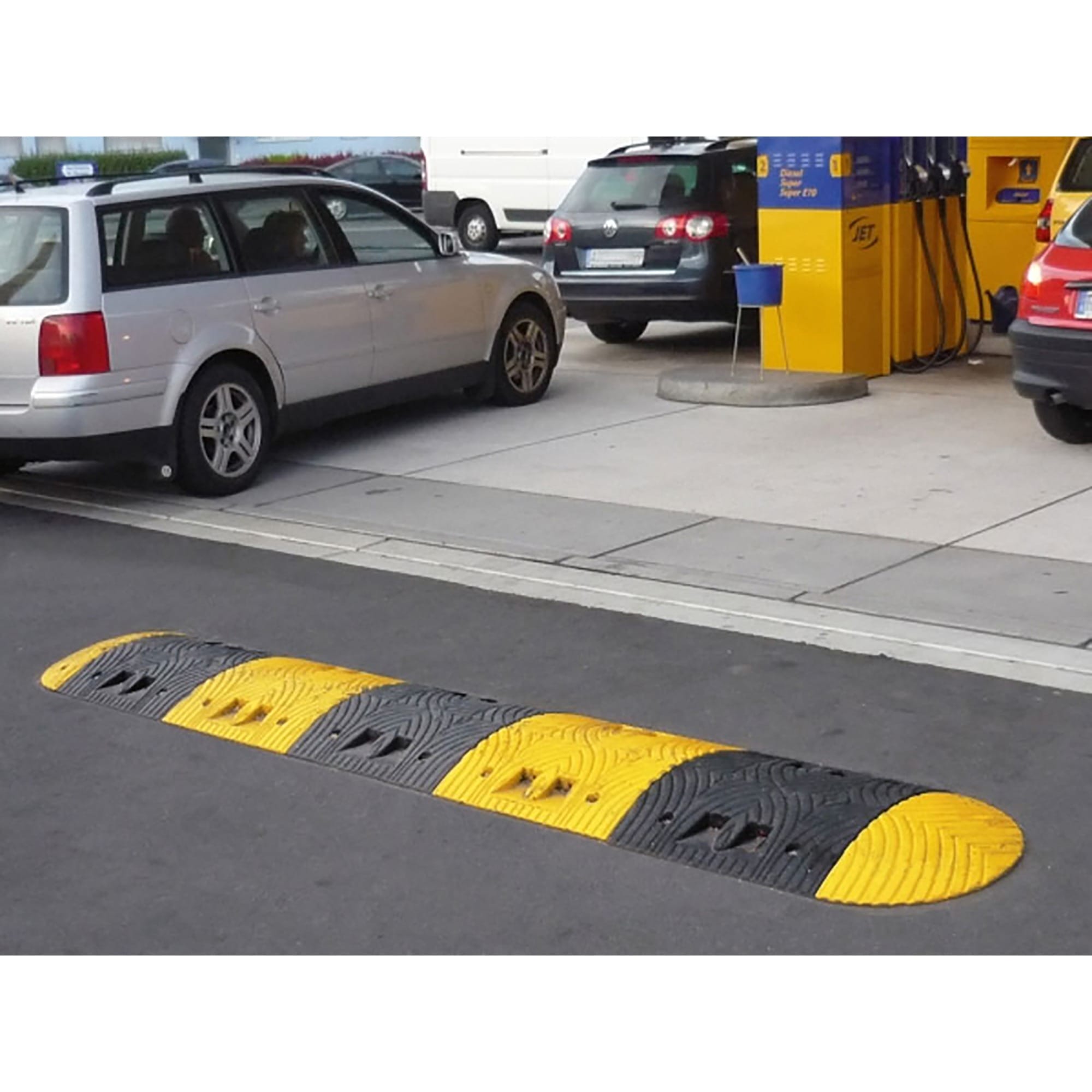 Schwarz-gelbe TOPSTOP-Fahrbahnschwelle auf Tankstellenpflaster, dahinter geparkte Autos und Zapfsäulen gut sichtbar.