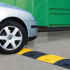 Vorderreifen eines silbernen Autos nähert sich gelb-schwarzer TOPSTOP Fahrbahnschwelle auf grauem Asphalt, grüne Struktur hinten.