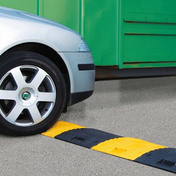 Vorderreifen eines silbernen Autos nähert sich gelb-schwarzer TOPSTOP Fahrbahnschwelle auf grauem Asphalt, grüne Struktur hinten.