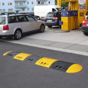 Gelb-schwarze TOPSTOP-Fahrbahnschwelle an Tankstelle neben Zapfsäulen, mehrere parkende Autos in der Nähe sichtbar.