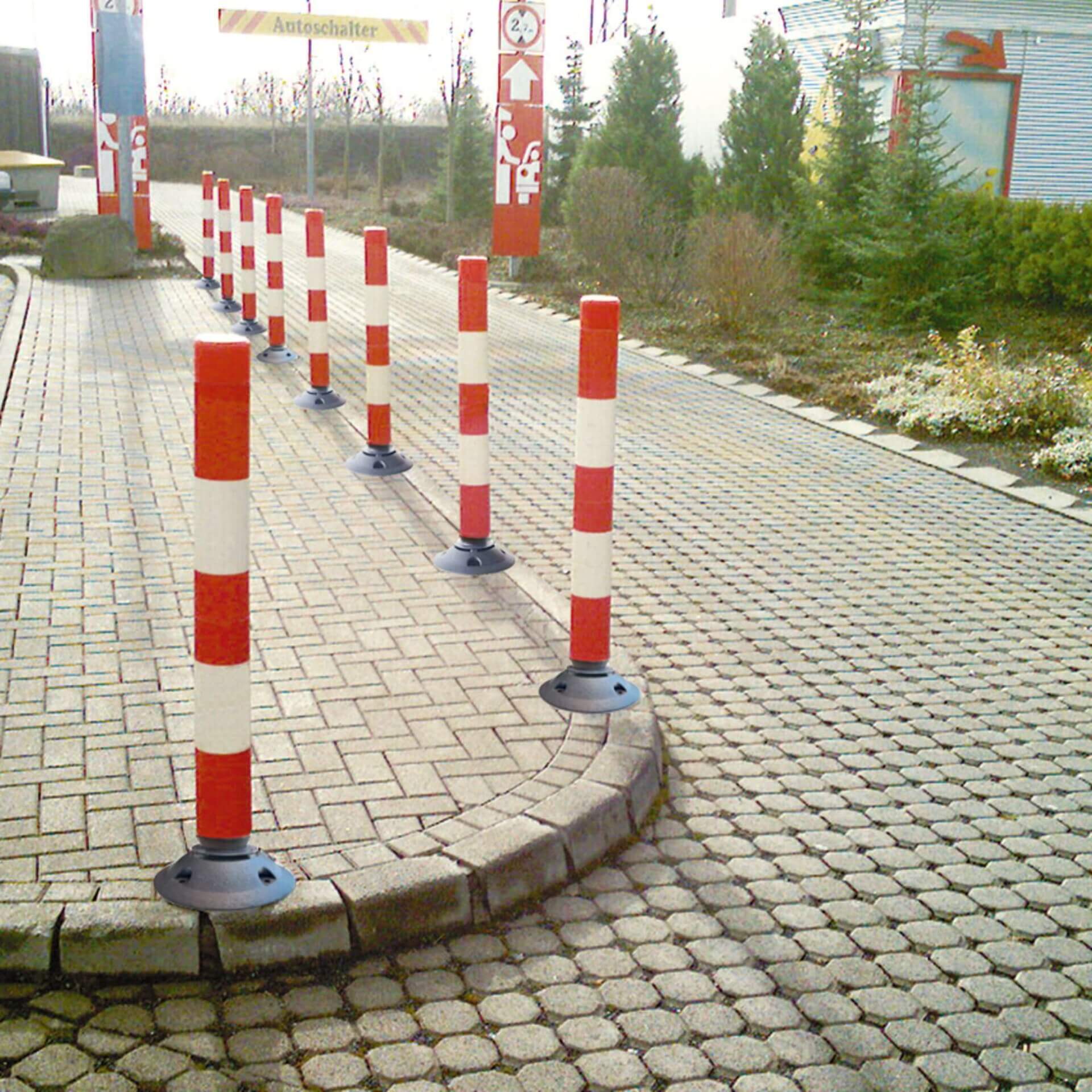 LeitPin rot-weiß gestreifte Poller säumen einen gebogenen, gepflasterten Weg neben Sträuchern und Bäumen im Landschaftsbereich.