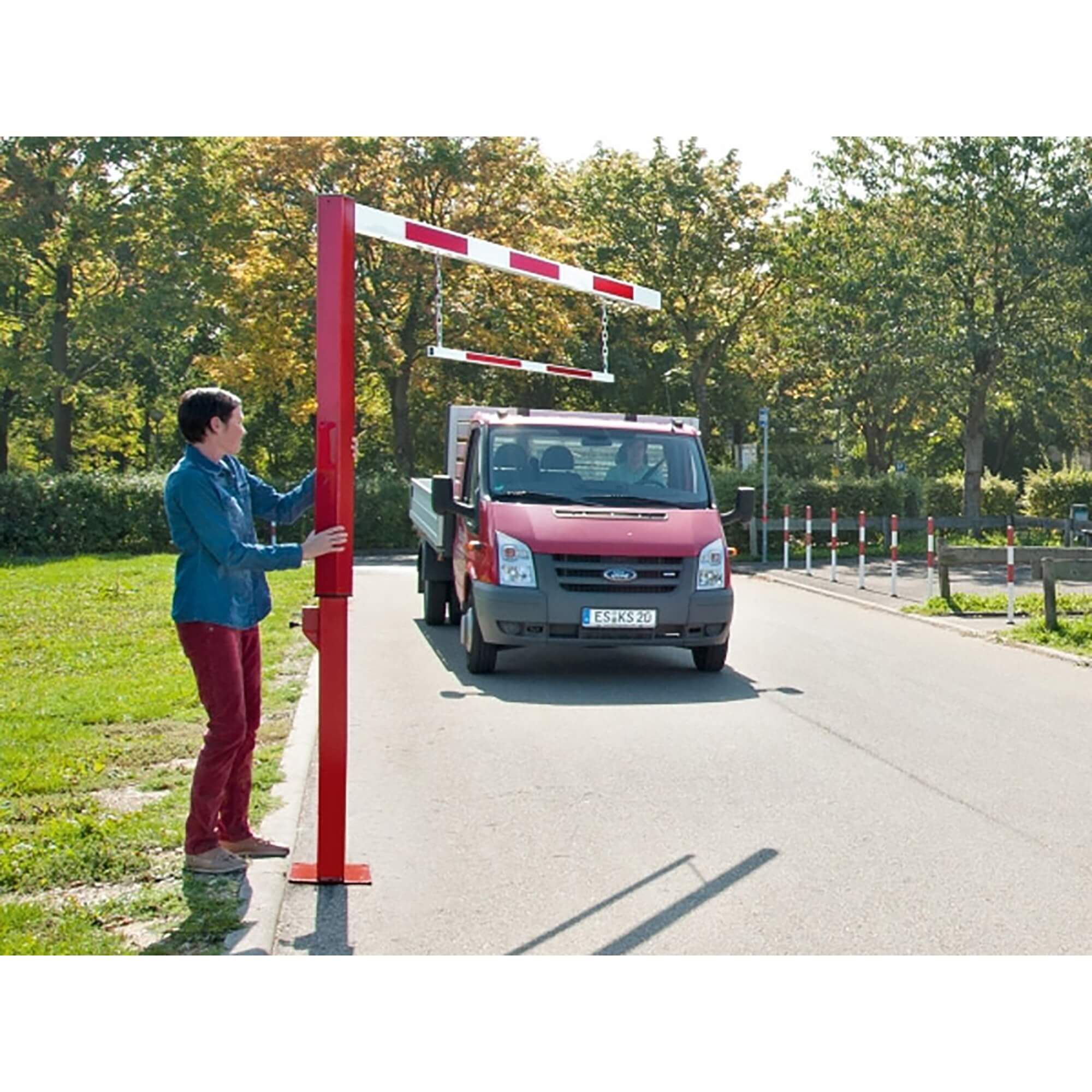 Eine Person stellt einen COMPACT-Durchfahrtbegrenzer drehbar ein, roter Nutzfahrzeug-LKW nähert sich auf Asphaltstraße im Grünen.