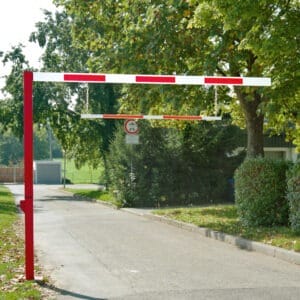 Rot-weißer Durchfahrtbegrenzer über geteerten Straße, seitlich Bäume und Büsche, Verkehrsschild für Höhenbegrenzung.