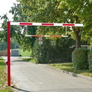 Rot-weißer Durchfahrtbegrenzer über geteerten Straße, seitlich Bäume und Büsche, Verkehrsschild für Höhenbegrenzung.
