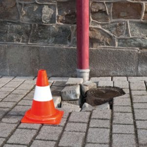 Reflektierendes Leitkegel auf Gehweg neben gebrochenen, unebenen Pflastersteinen, Abflussrohr und Kanaldeckel.