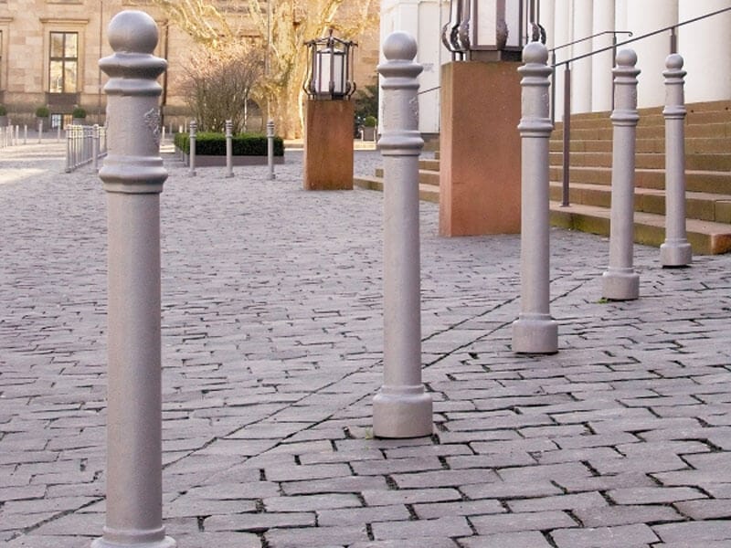 Kurze, weiße Wappen-Poller sichern einen gepflasterten Gehweg neben einem Gebäude mit Treppe und Außenlaternen.