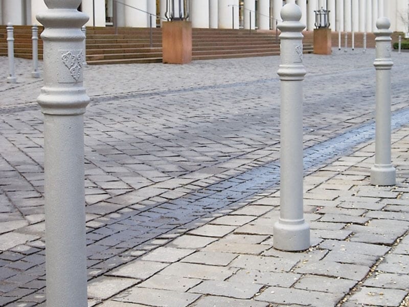 Reihe grauer Wappen-Poller auf Kopfsteinpflaster vor Gebäude mit Treppenstufen und großen Säulen, Barrierefreiheit.