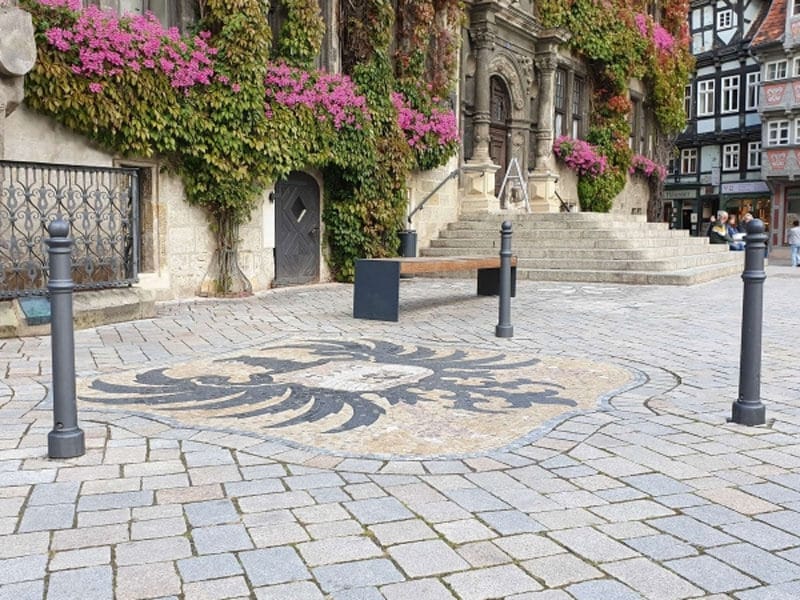 Wappen-Poller auf Kopfsteinpflaster mit großem Adler, umgeben von Pollern, vor historischem Gebäude und efeubewachsenen Mauern.